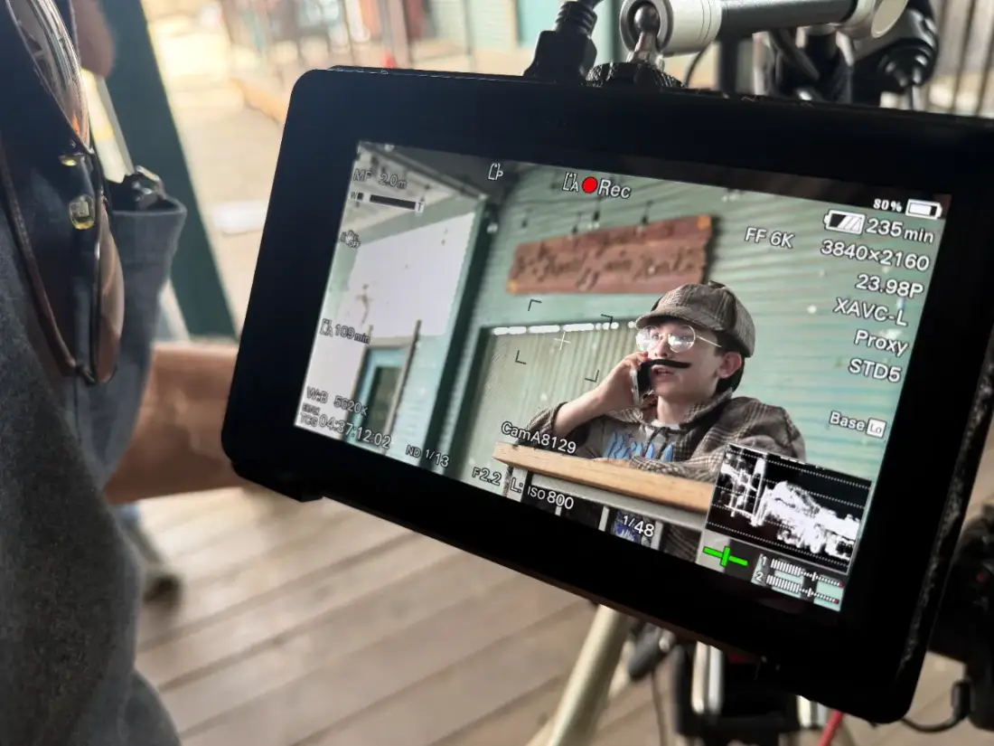 A young actor in a flat cap and moustache framed up on the camera monitor, mid-take