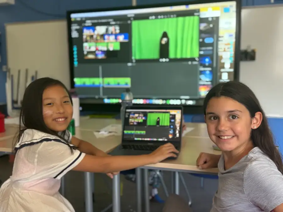 Two campers cutting their green-screen footage on a laptop, the timeline up on the big screen